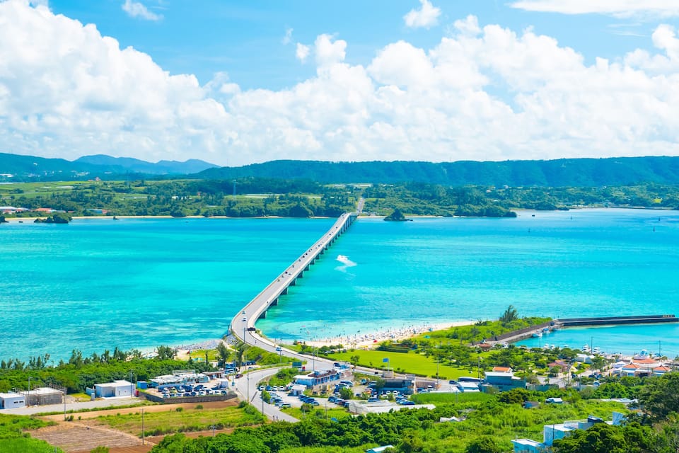 A coastal town with a long pier extending into turquoise waters, surrounded by lush greenery and mountains.