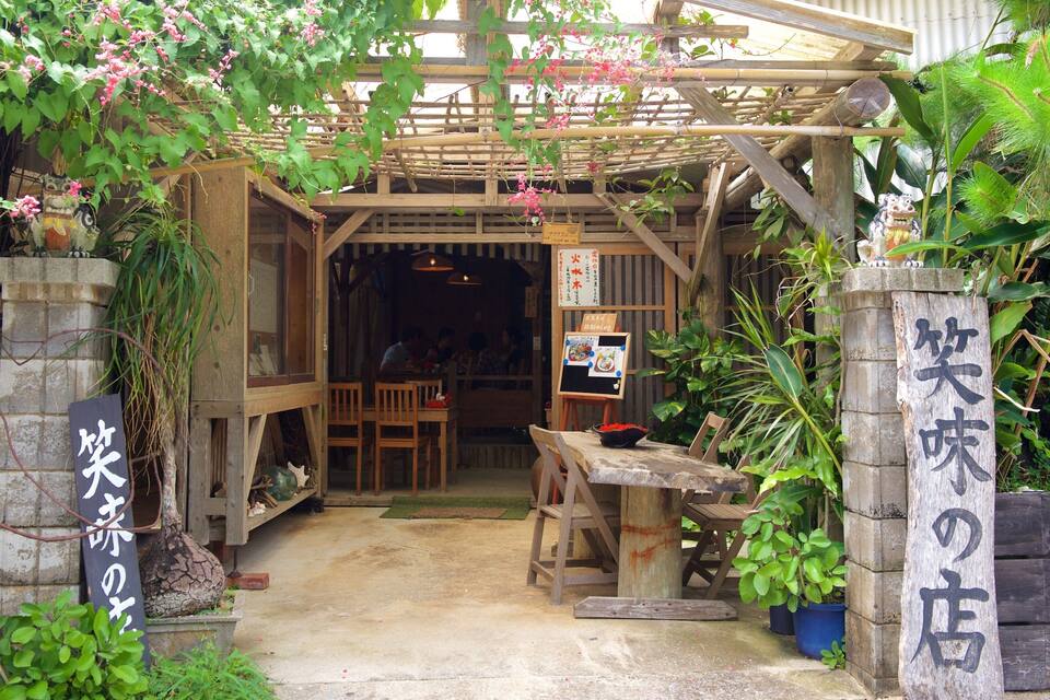 A rustic wooden entrance with a bamboo roof, surrounded by lush greenery and potted plants.