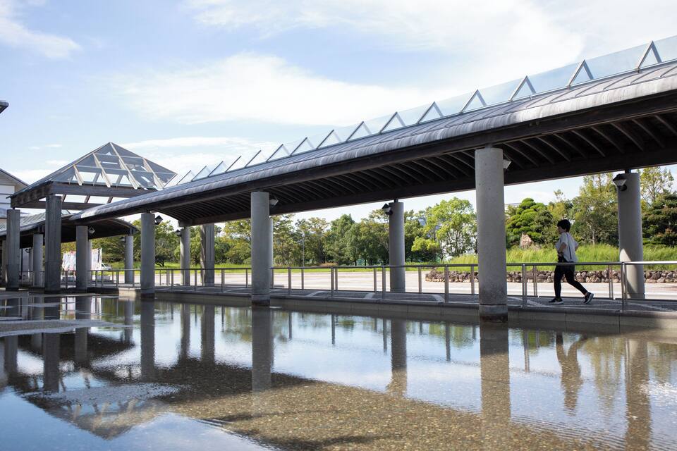 A pedestrian bridge over a reflective water body with a geometric glass structure in the background.