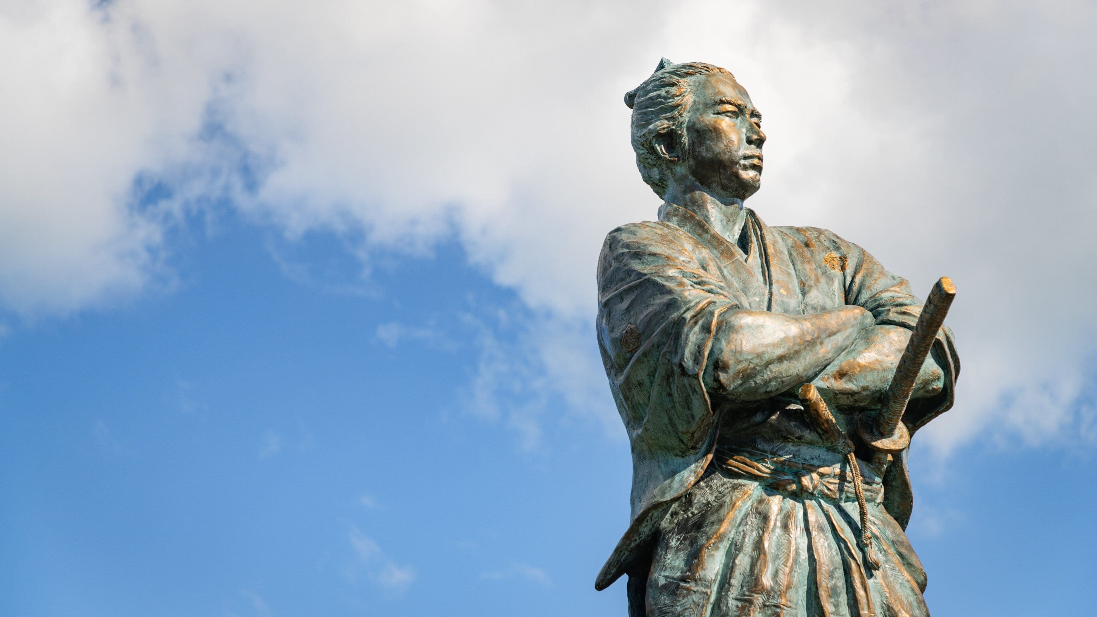 Eine Statue eines Mannes in traditioneller Kleidung mit verschränkten Armen vor blauem Himmel mit Wolken.