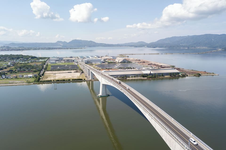 A large bridge spanning a wide river with industrial buildings and a distant mountain range.