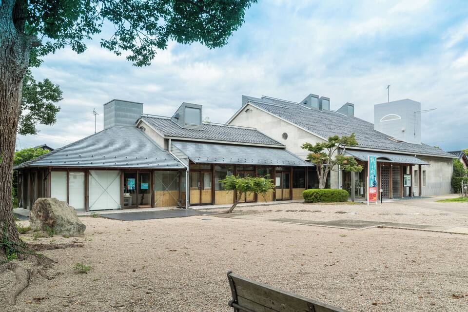 Um edifício tradicional japonês, com telhado de telhas e estrutura de madeira, rodeado por cascalho e árvores.
