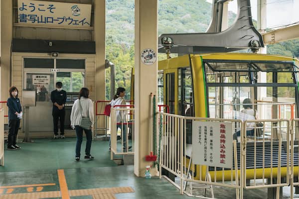 A yellow cable car station with people waiting, a sign with Japanese text, and a mountainous landscape in the background.