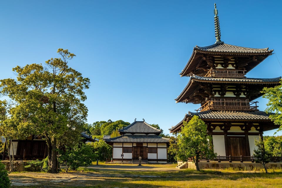 A traditional Japanese pagoda with multiple tiers, surrounded by lush greenery and a smaller building in the background.