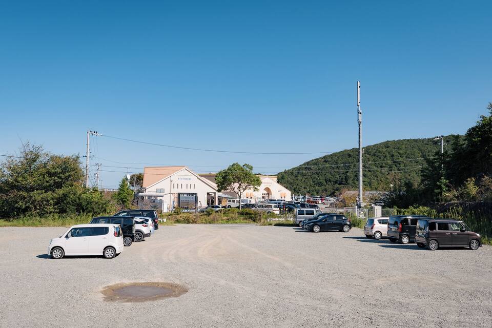 A gravel parking lot with several cars, a building with a red roof, and a hill with trees in the background.
