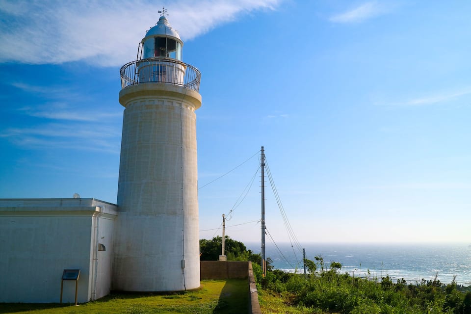 Un faro con torre blanca, barandilla de metal y zona verde a su lado.