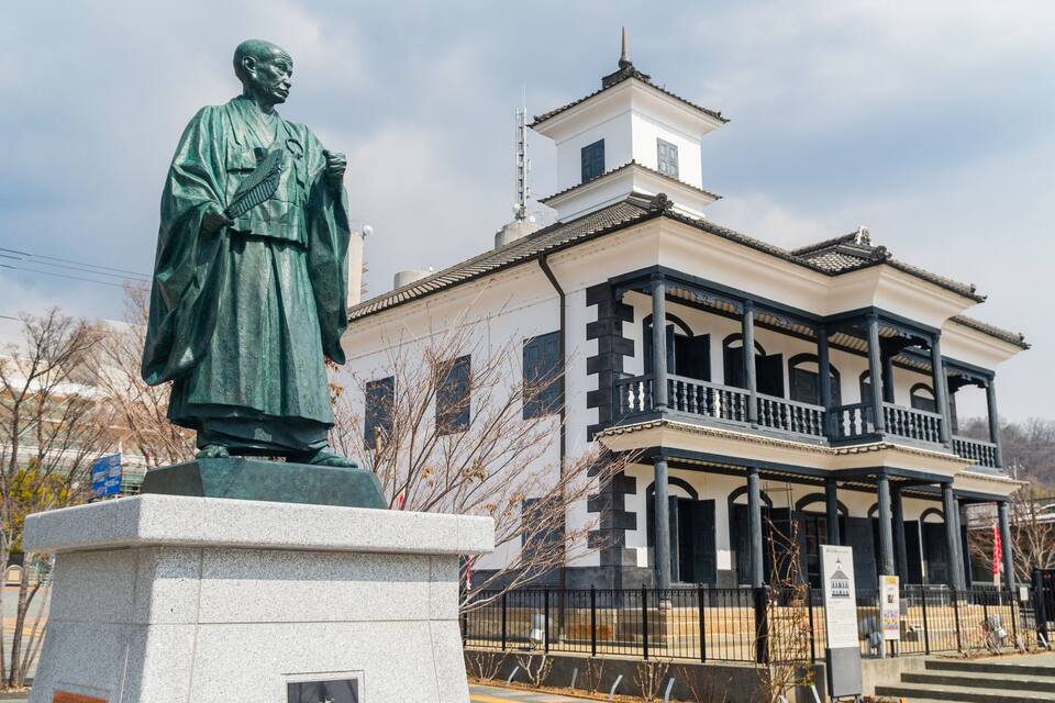 A statue of a historical figure in traditional attire, a traditional building with a balcony, and modern high-rise buildings in the background.