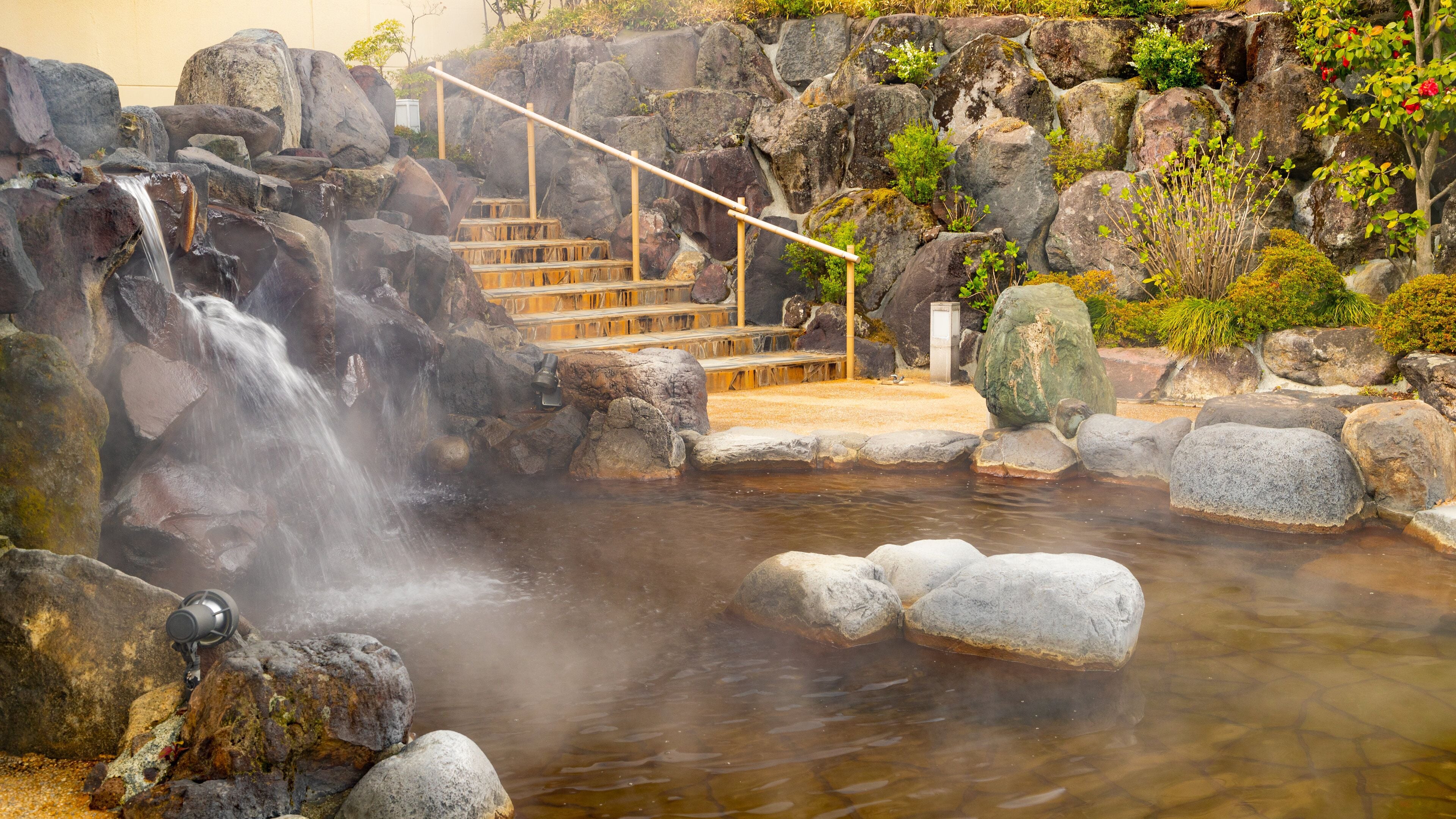 Perinteinen japanilainen onsen-kylpylä, johon johtaa puinen portaikko. Kylpylä sijaitsee kuuman lähteen äärellä, ja sen ympärillä on kiviä ja vehreää kasvillisuutta.