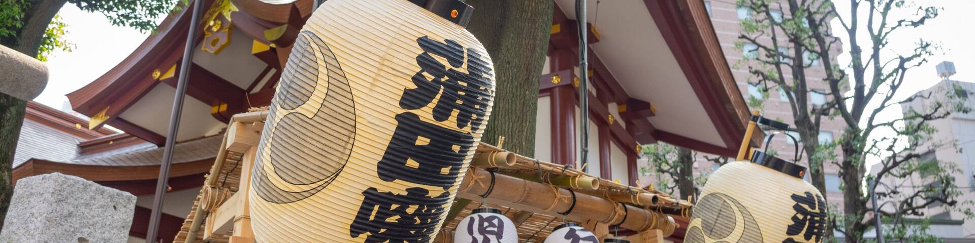 Traditional Japanese lanterns with calligraphy, a wooden structure, and a building with a red roof in the background.