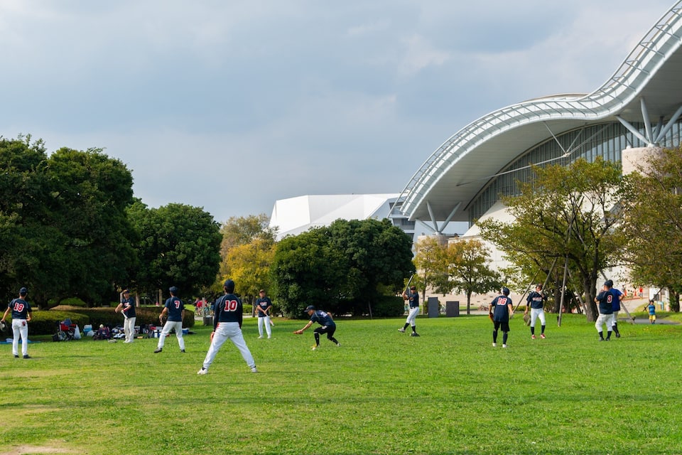 En baseballplan med spelare i matchställ, träd och en modern byggnad i bakgrunden.