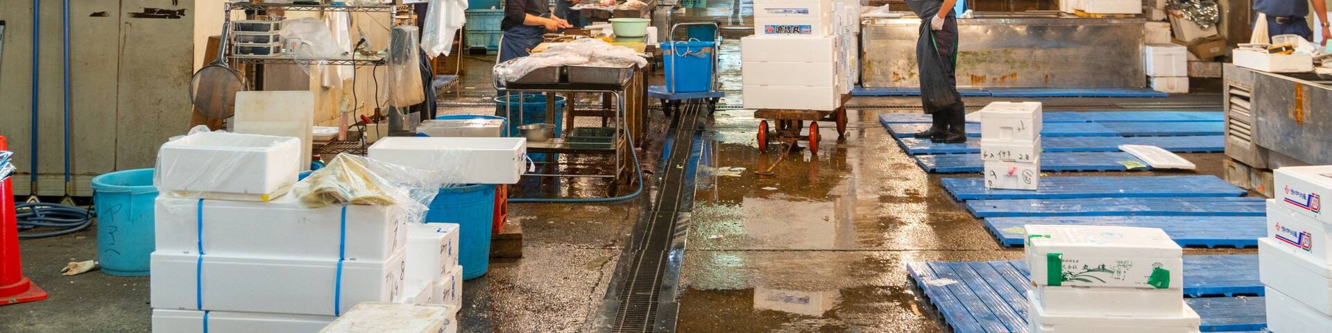 A busy fish market with workers handling fish, tables with fish boxes, and signs in a foreign language.