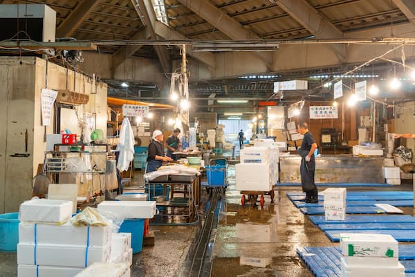 Un marché aux poissons animé, où l’on voit des employés manipuler du poisson, des tables remplies de caisses de poissons et des panneaux affichés dans une langue étrangère.