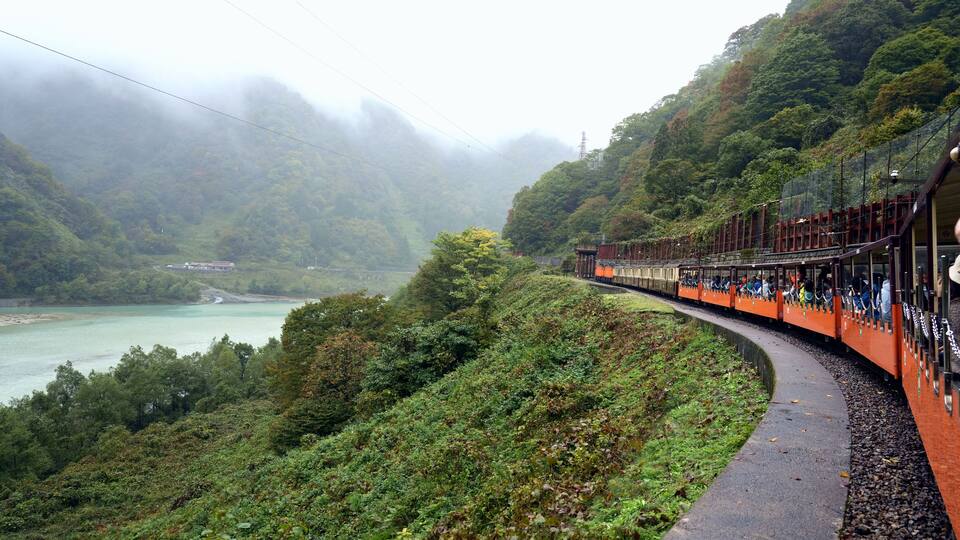 A train on a curved track through a mountainous area with a river and dense greenery.