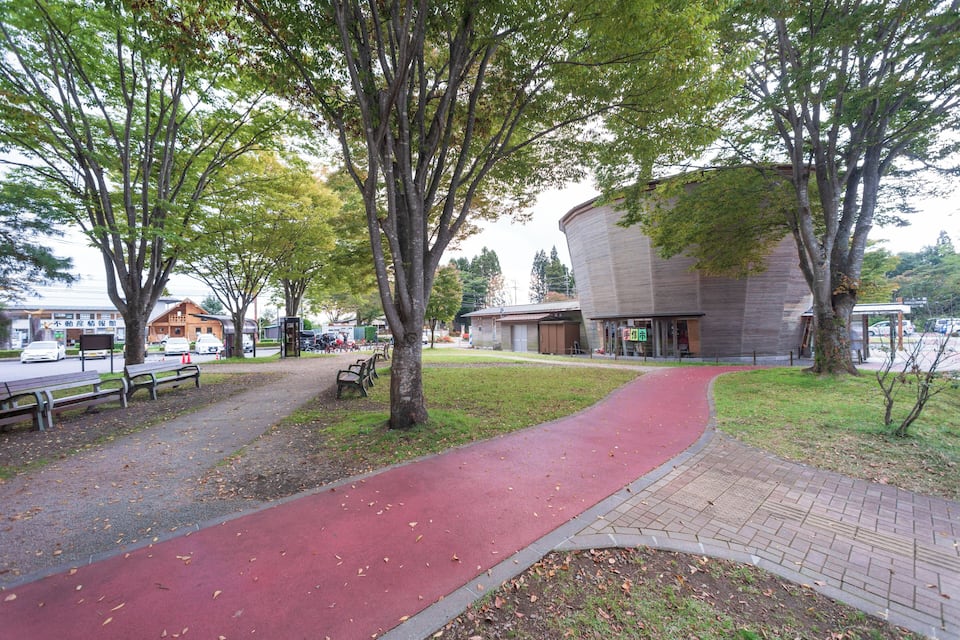 Een park met een rode fietspad, bankjes, bomen en een gebouw met een opvallend gebogen dak.