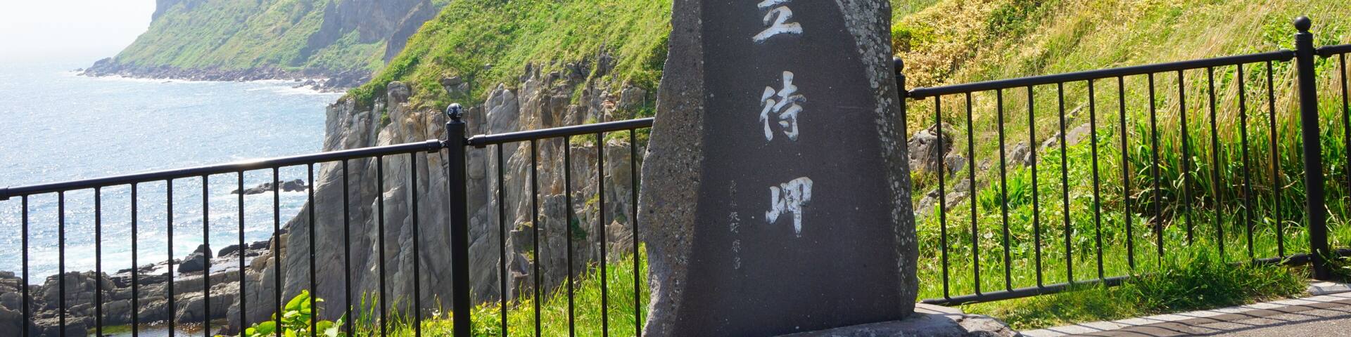A coastal landscape with a stone marker, a metal fence, and a steep cliff.