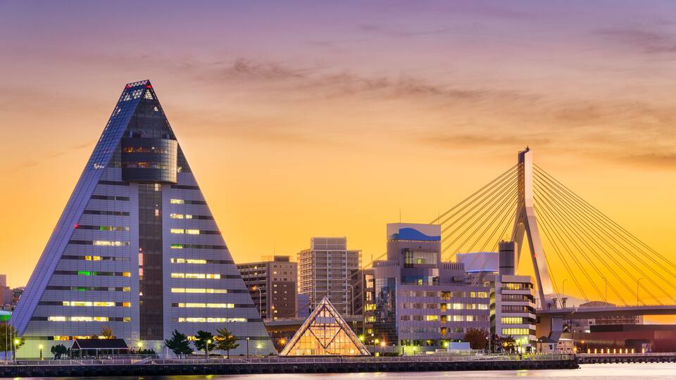 A cityscape with a prominent triangular building and a cable-stayed bridge at sunset.