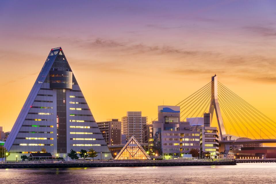 A cityscape with a prominent triangular building and a cable-stayed bridge at sunset.