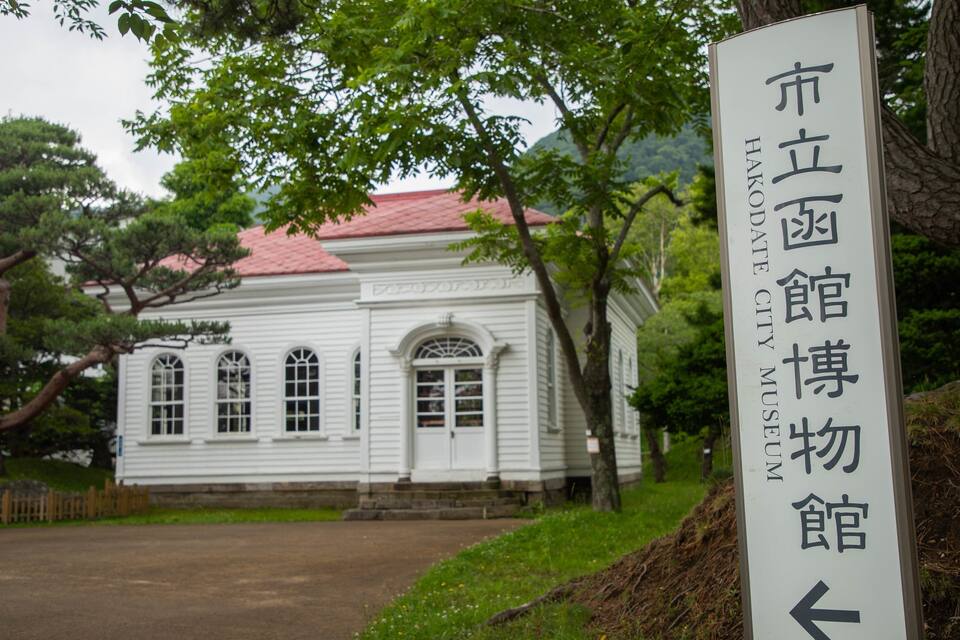 Ein traditionelles Gebäude mit rotem Dach und weißen Wänden, umgeben von Bäumen. Ein Schild mit japanischen Schriftzeichen zeigt nach links.