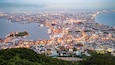 A coastal cityscape with a harbor, dense urban area, and a prominent hill with greenery.