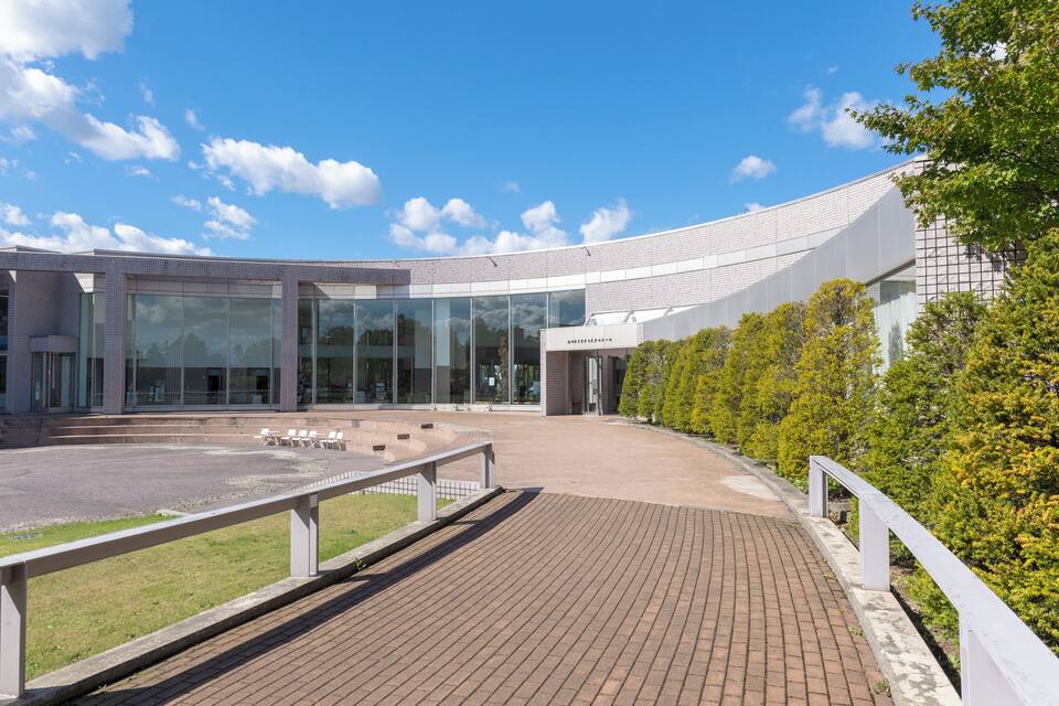 Un edificio moderno con amplios ventanales, techo curvo y un sendero pavimentado con barandillas blancas.