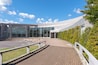 A modern building with large glass windows, a curved roof, and a paved walkway with white railings.