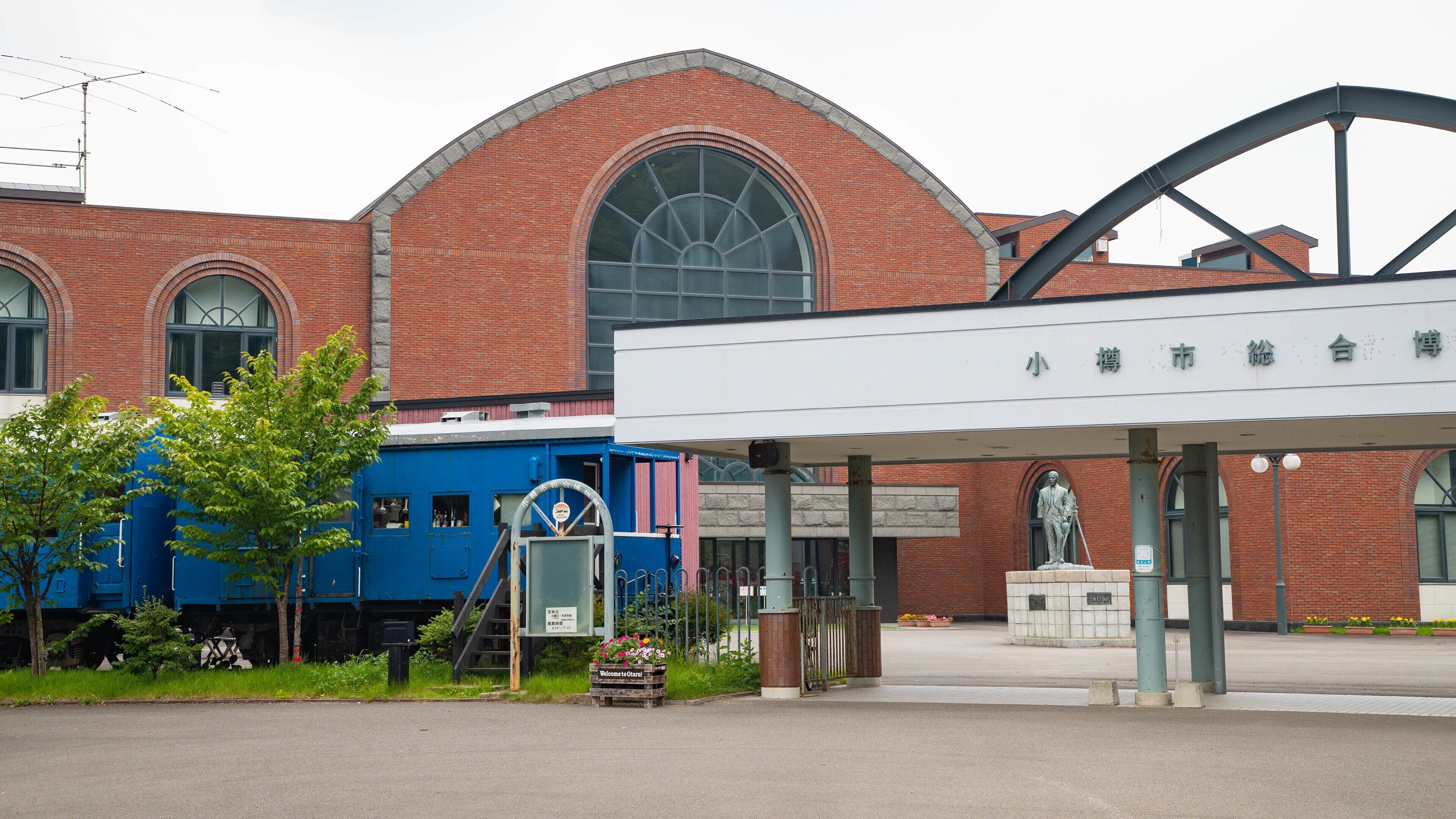 A train station with a blue locomotive, a statue, and a brick building with arched windows.
