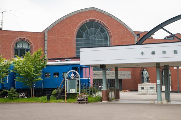 A train station with a blue locomotive, a statue, and a brick building with arched windows.