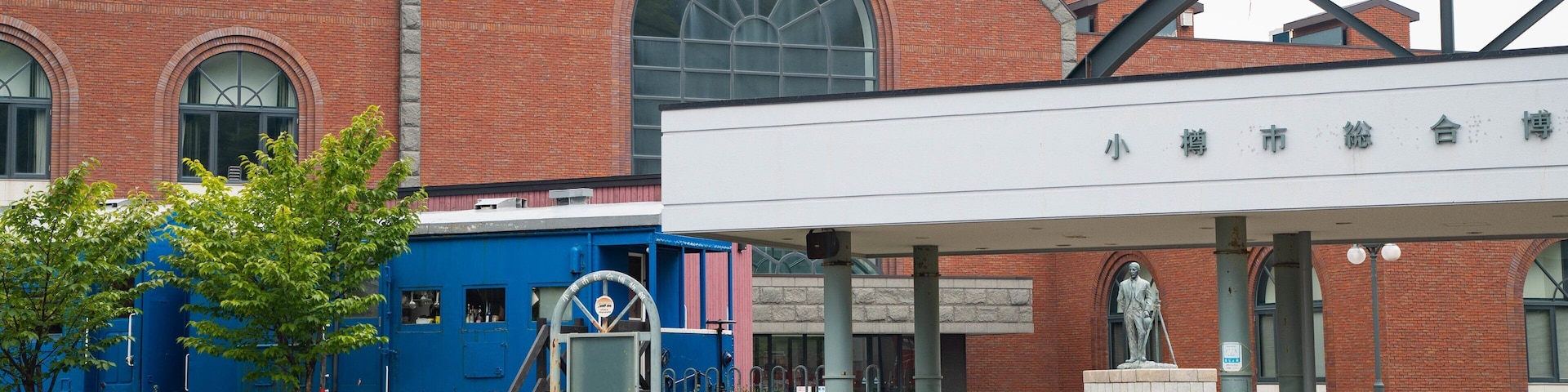 Uma estação de trem com uma locomotiva azul, uma estátua e um edifício de tijolos com janelas em arco.