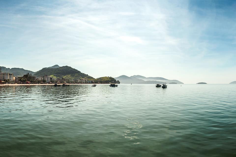 Ruhiges Meer mit Booten, einem Strand und Bergen im Hintergrund.