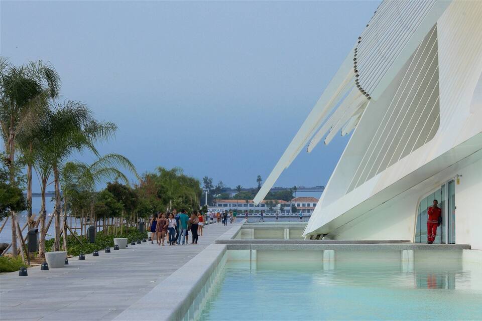 A modern building with a pool and people walking along a waterfront promenade.