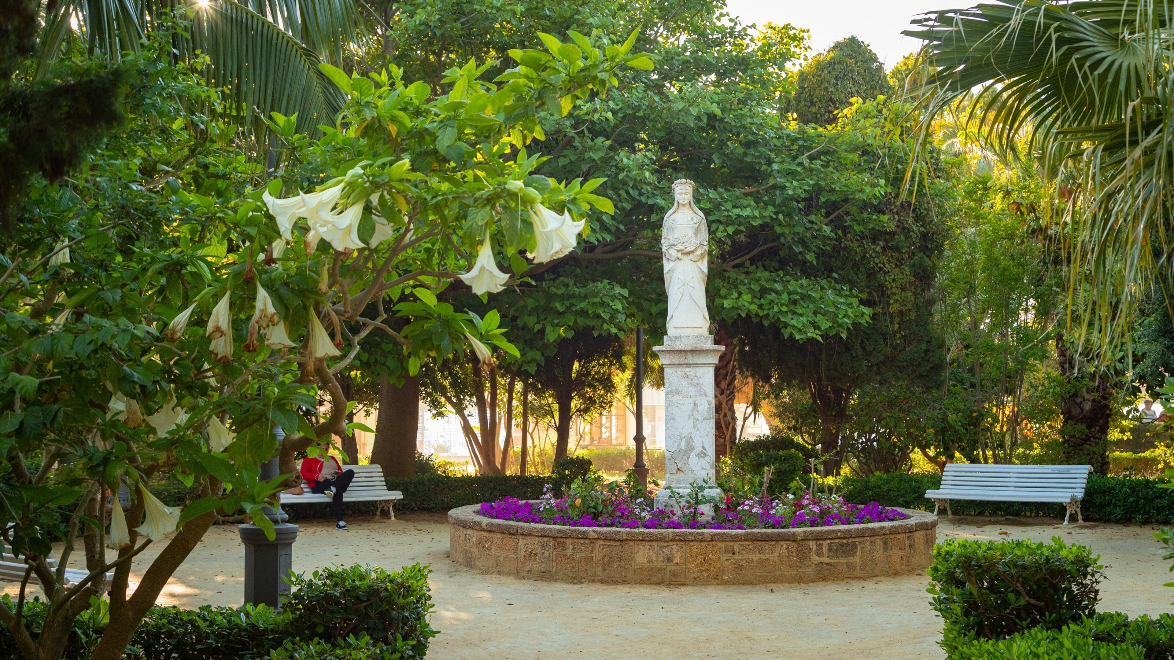 A statue in a circular flower bed surrounded by lush greenery and benches.