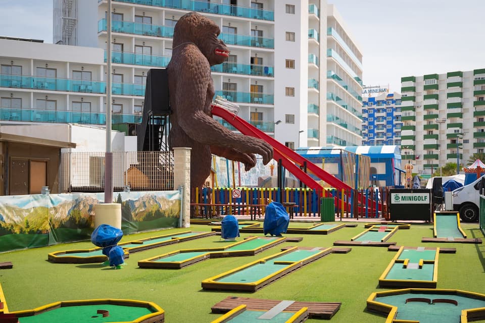 A mini-golf course with a large gorilla statue, a slide, and a building with balconies in the background.