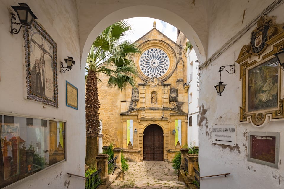 Ein gewölbter Gang führt zu einer historischen Kirche mit einem großen Rundfenster und einer Palme.