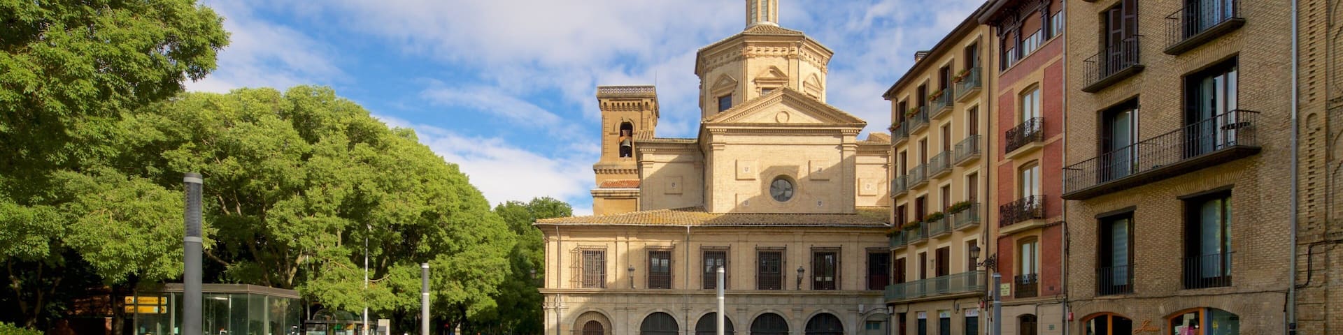 A historic church with a dome and bell tower, surrounded by lush trees and a well-maintained square with benches.