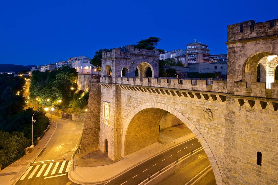 Eine Stebrücke mit Bögen, eine Straße darunter und Gebäude im Hintergrund.