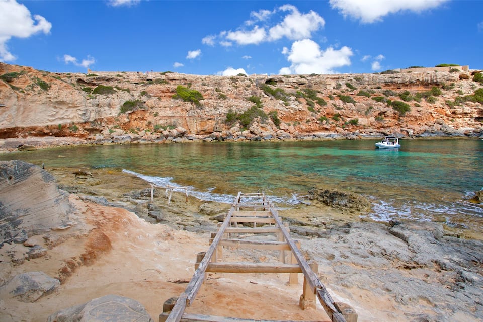 A rocky beach with a wooden ladder leading into clear turquoise waters, a boat on the water, and a cliff with sparse vegetation.
