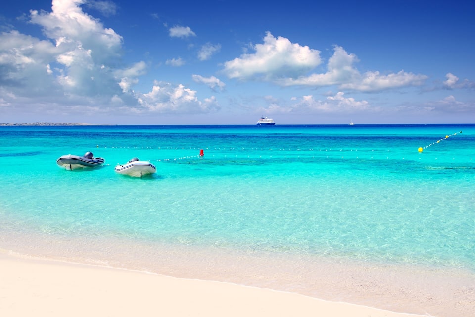 A beach with clear turquoise water, two small boats, a large cruise ship in the distance, and a sandy shore.