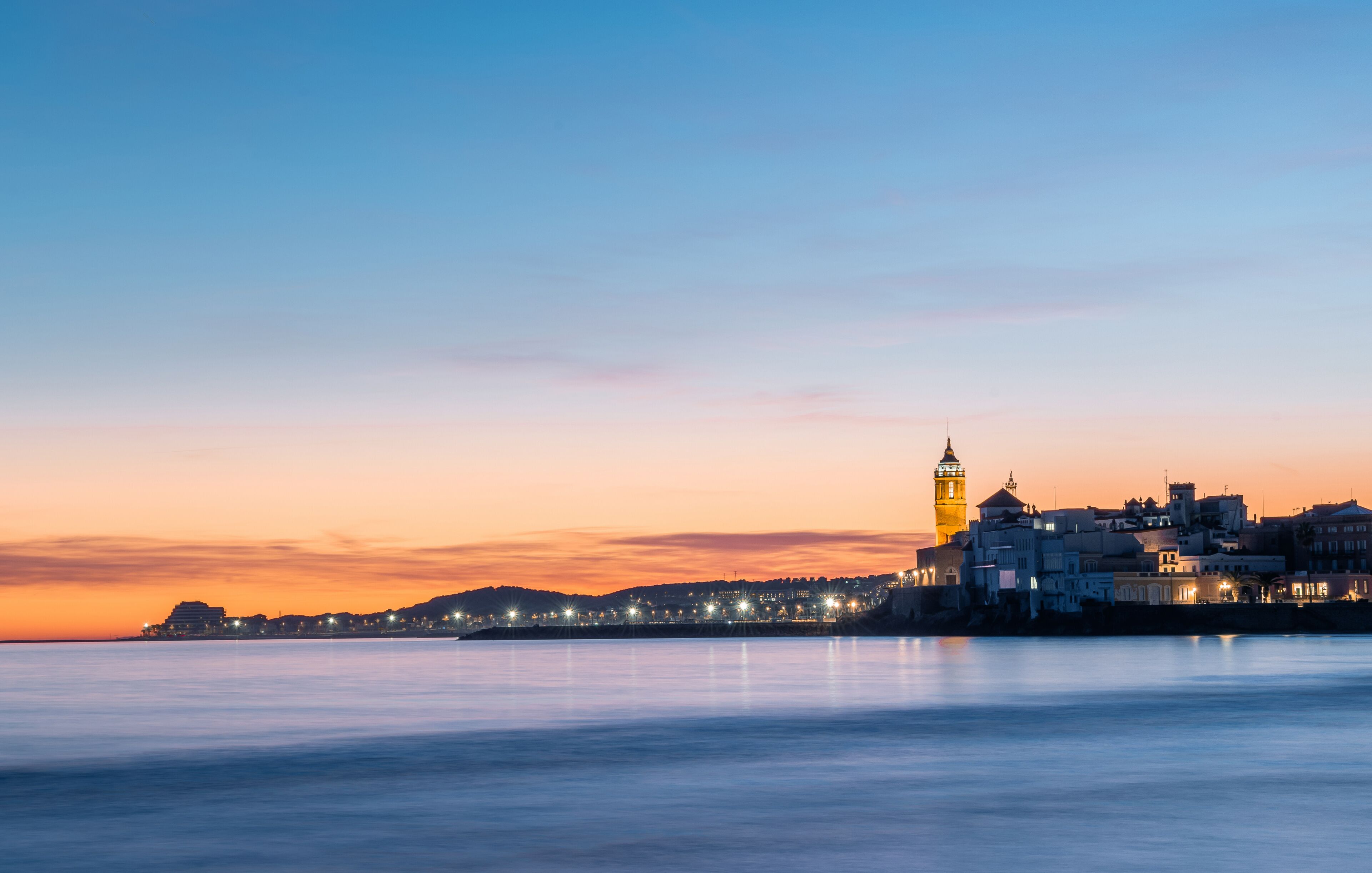 Fotografia de larga exposición de Sitges desde la playa con la iglesia iluminada al fondo en un atardecer, anochecer con cielo de colores azul, naranja y amarillo y unas ligeras nubes en el horizonte