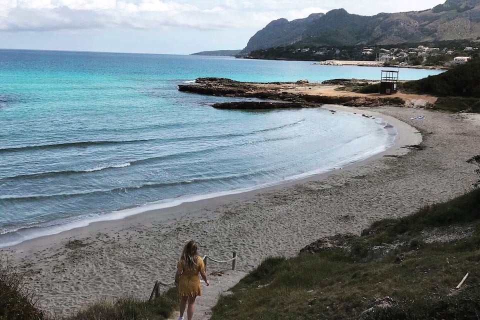 Un paysage côtier avec une plage de sable, un promontoire rocheux et une personne qui marche le long d’un sentier.