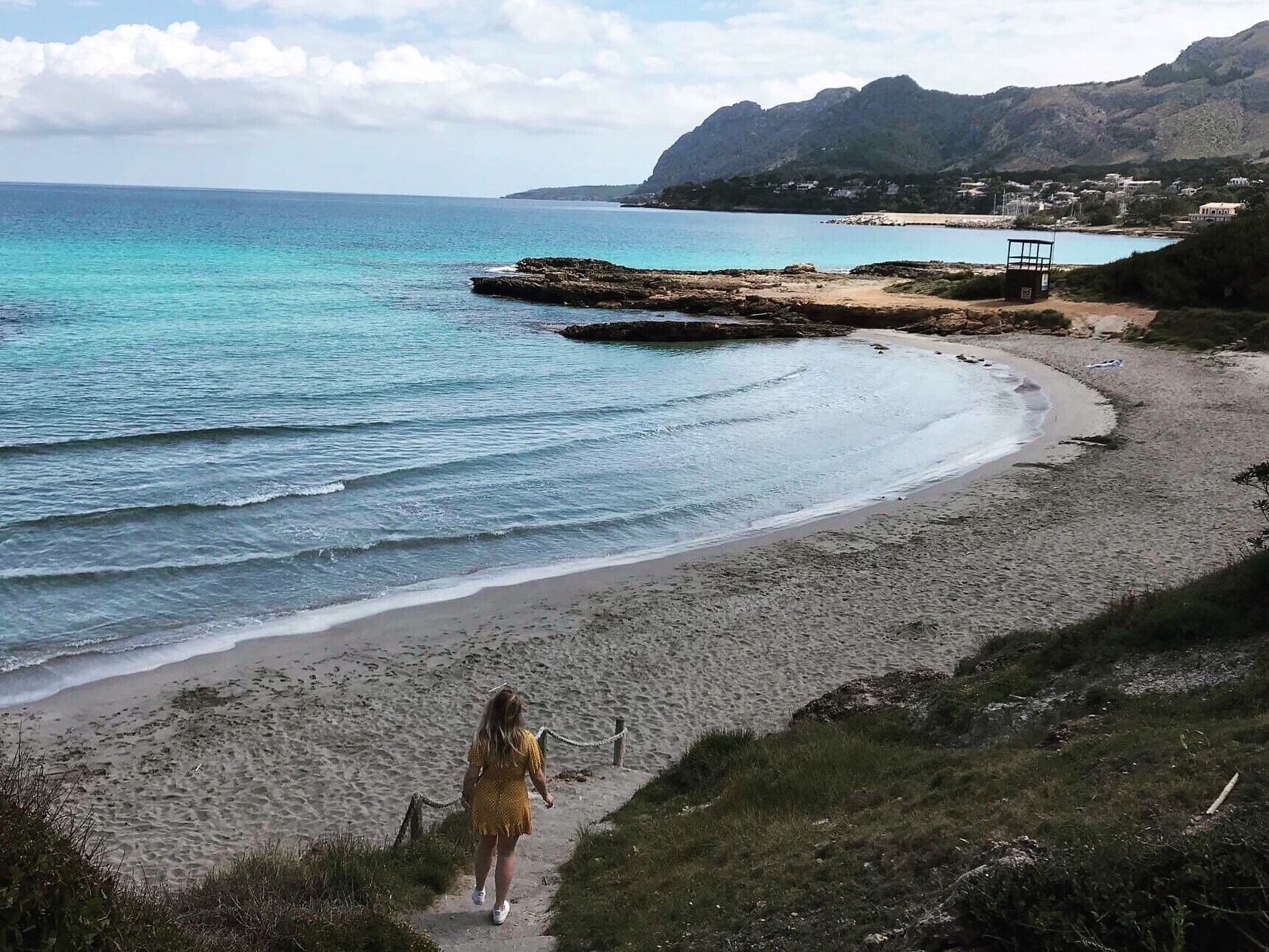 A coastal landscape with a sandy beach, a rocky outcrop, and a person walking along a path.