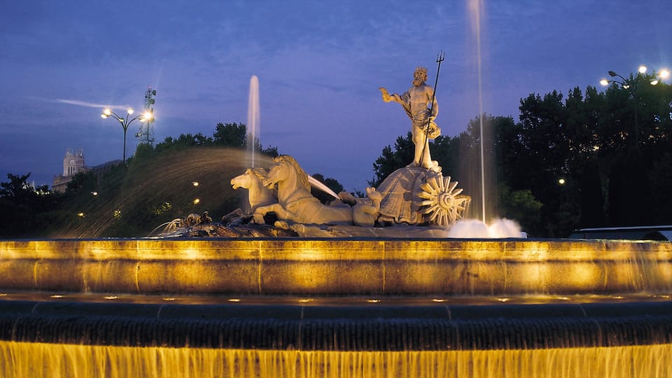 Une fontaine illuminée la nuit, ornée d’une statue représentant un homme tenant un trident et un cheval.