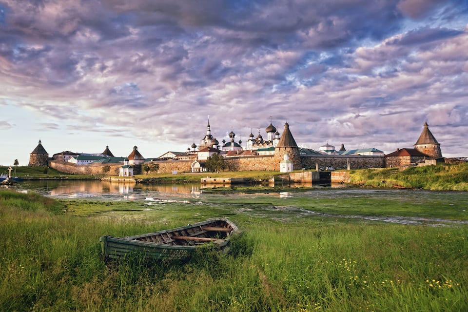 A historic fortress with multiple domed towers, a river in the foreground, and a small boat.