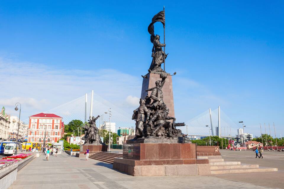 Una gran escultura al aire libre con una bandera, un puente al fondo y un edificio de techo rojo.
