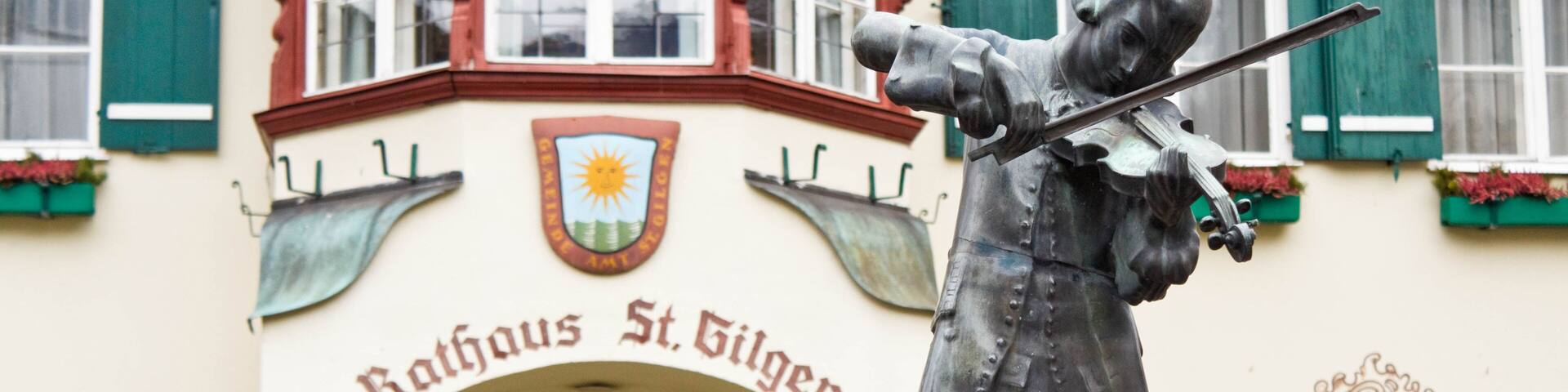 A statue of a man playing a violin in front of a building with green shutters and a coat of arms.