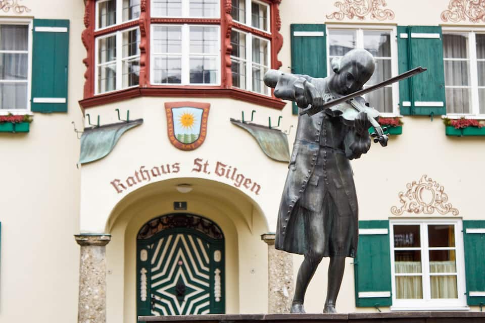A statue of a man playing a violin in front of a building with green shutters and a coat of arms.
