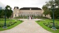 A grand building with a symmetrical facade, a central staircase, and a well-maintained garden with a gravel path leading to the entrance.