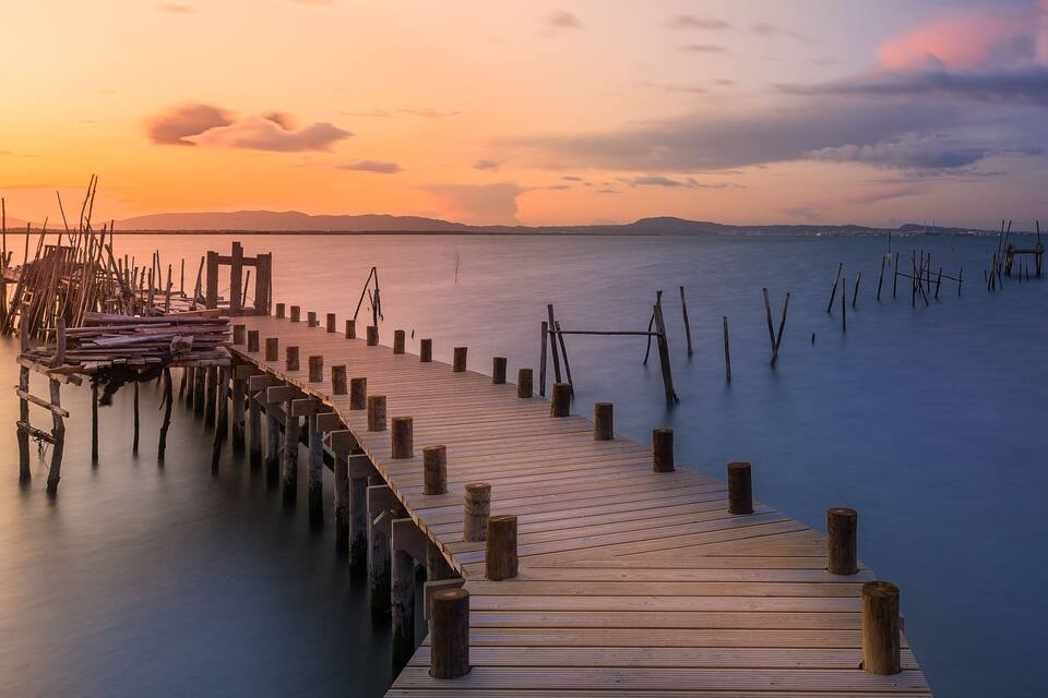 A wooden pier extending into calm waters at sunset with distant mountains and a clear sky.