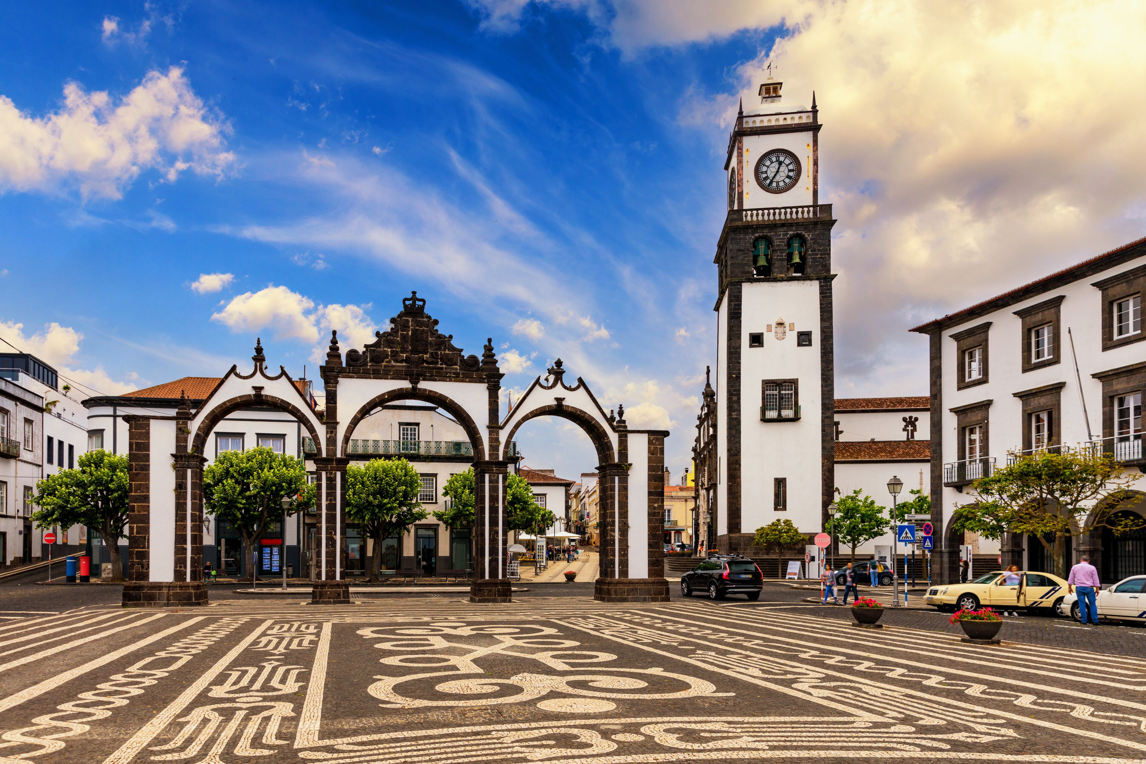 A historic plaza with a prominent clock tower, arches, and a patterned pavement.