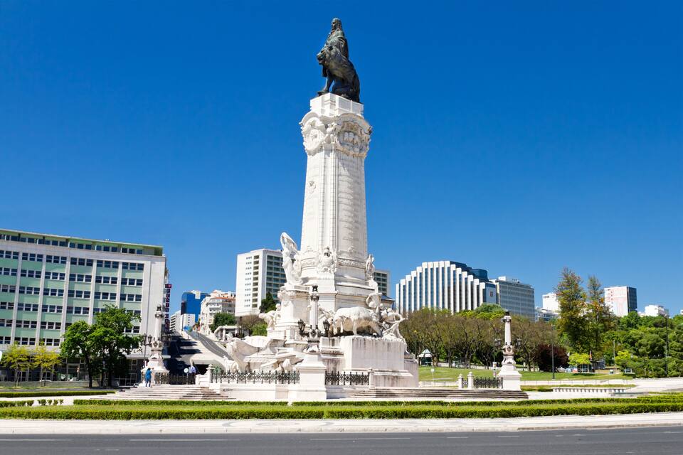 Um monumento alto com uma estátua no topo, rodeado por um parque bem cuidado com árvores e um céu azul límpido.