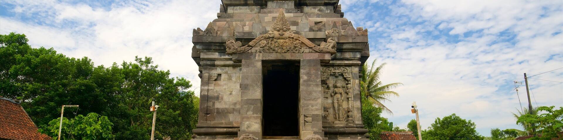 A stone temple with a central doorway, flanked by two large stone lions, set against a backdrop of lush greenery and a partly cloudy sky.